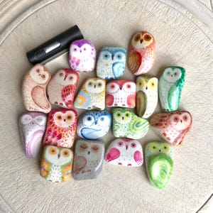 May include: A collection of hand-painted owl figurines in various colors and designs. The owls are painted on smooth, rounded stones, with details like feathers, eyes, and beaks. The color palette includes shades of pink, blue, green, and orange.