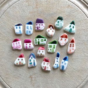 May include: A collection of 17 small, hand-painted ceramic houses in various colours and designs. Each house is unique and features different details, such as windows, doors, and chimneys. The houses are arranged in a circle on a white surface.