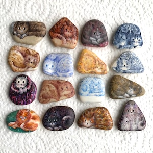 May include: A collection of 16 painted stones featuring various cat designs. The stones are painted in a variety of colours, including brown, black, white, blue, and grey. The cats are depicted in different poses, including sleeping, sitting, and standing.