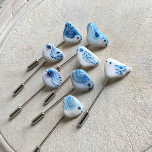 May include: Eight bird-shaped lapel pins. Each pin features a small, white ceramic bird with blue painted details. The birds are mounted on long, silver-coloured metal pins with a decorative cap at the end.