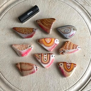 May include: Nine hand-painted ceramic birds with different designs. Each bird is painted with a variety of colors, including pink, orange, yellow, brown, and white. The birds are all different shapes and sizes, and they are all facing the viewer.