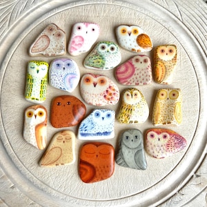 May include: A selection of hand-painted owl ornaments in diverse colours and patterns. The owls are arranged on a light-coloured, textured surface. Each owl features unique details, including different eye shapes and wing designs.