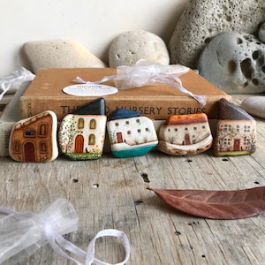 May include: Six hand-painted miniature houses on rocks, each with a unique design and color scheme. The houses are arranged in a row on a wooden surface. The houses are painted in shades of brown, white, blue, and green. The houses have small windows and doors, and some have flowers and trees painted around them.