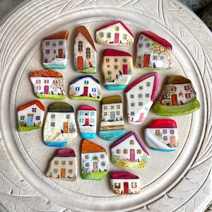 May include: A selection of hand-painted miniature house-shaped stones. Each stone has a unique design with colourful roofs, doors, windows, and some include small black cat figures. The houses are arranged on a decorative, off-white surface.
