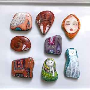 May include: Eight painted stones with various designs, including a fox, a sleeping fox, a woman's face, two elephants, and two cats. The stones are on a white surface.