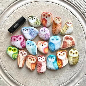 May include: A selection of hand-painted owl ornaments in diverse colours and patterns. The owls are in various sizes and feature shades of pink, blue, green, and orange. A black lighter is in the upper left corner.