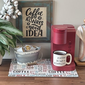 May include: A red Keurig coffee maker with a coffee mug that reads "Coffee Books & Mountains" on a countertop with a coffee-themed mat. A sign above the coffee maker reads "Coffee is always a good idea".