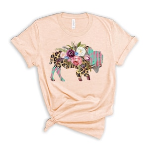 May include: A light peach t-shirt with a floral design on a buffalo silhouette. The buffalo is covered in a leopard print and has a turquoise and pink watercolor background.