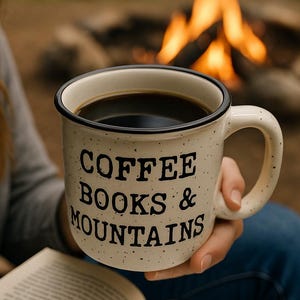 May include: A speckled white coffee mug filled with dark coffee, with the text "COFFEE BOOKS & MOUNTAINS" in black. The mug has a black rim and handle. A campfire is visible in the background.