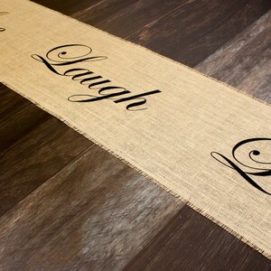 May include: A burlap table runner with the words "Live, Laugh, Love" printed in black cursive font.