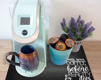 May include: A black chalkboard coffee mat with the text "May your coffee kick in before reality does". A teal blue mug, a light blue coffee maker, and a teal blue container filled with coffee pods are on the mat.