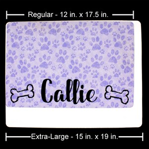 May include: A purple and white dog food mat with a paw print pattern. The mat has the name "Callie" printed on it with two bone shapes on either side of the name. The mat is available in two sizes: 12 inches by 17.5 inches and 15 inches by 19 inches.