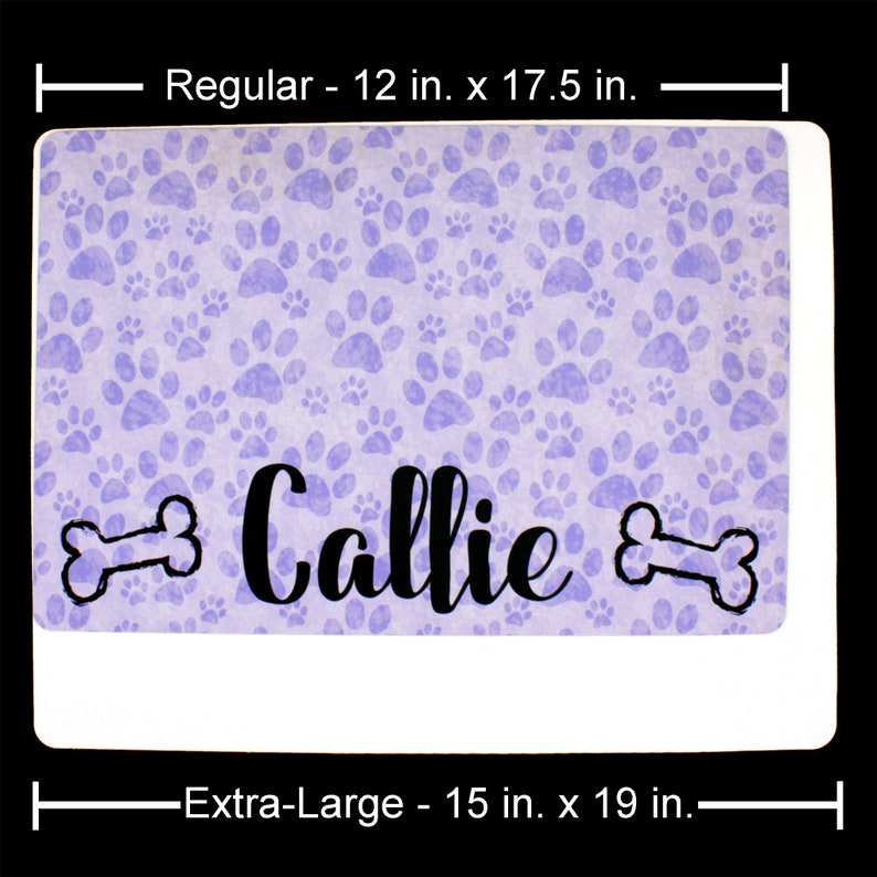 May include: A purple and white dog food mat with a paw print pattern. The mat has the name "Callie" printed on it with two bone shapes on either side of the name. The mat is available in two sizes: 12 inches by 17.5 inches and 15 inches by 19 inches.