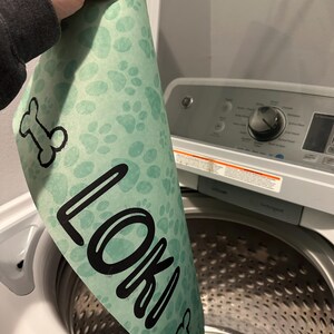 May include: A green dog bed with paw prints and a bone design. The bed has the name "Loki" printed on it. The bed is being placed in a washing machine.