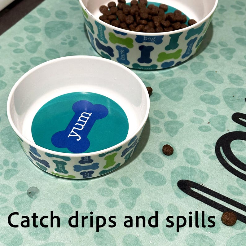 May include: Two white dog bowls with blue and green paw print designs. The bowls are on a green mat with paw prints. One bowl has the word "yum" on it. The mat has the text "Catch drips and spills" on it.