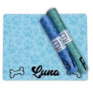 May include: A blue and green dog food mat with a paw print design. The mat has the name "Luna" written on it in black cursive font. Two rolls of blue and green paper are on top of the mat. The paper is labeled "Paw Print" and "Chewmat".