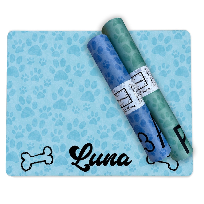 May include: A blue and green dog food mat with a paw print design. The mat has the name "Luna" written on it in black cursive font. Two rolls of blue and green paper are on top of the mat. The paper is labeled "Paw Print" and "Chewmat".