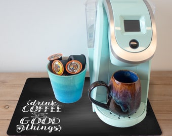 May include: A black chalkboard mat with the text "drink coffee and do good things" in white. A blue coffee maker with a silver trim and a brown ceramic mug are on the mat. A blue ceramic cup with coffee pods is on the mat.
