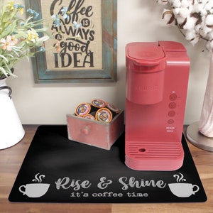 Puede incluir: Alfombra de cocina de pizarra negra con el texto "Rise & Shine it's coffee time" y una imagen estilizada de una taza de café con vapor. Una cafetera Keurig roja está sobre la alfombra.