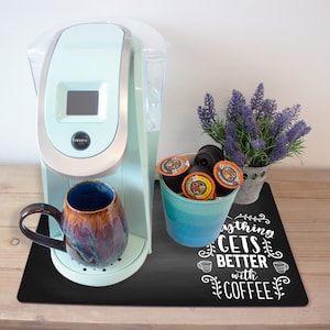 Può includere: Un tappetino da caffè nero con la scritta "Tutto diventa migliore con il caffè" in lettere bianche. Una macchina da caffè blu chiaro con una tazza marrone e un contenitore blu di capsule di caffè sono sul tappetino.