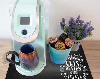 May include: A black coffee mat with the text "Everything gets better with coffee" in white lettering. A light blue coffee maker with a brown mug and a blue container of coffee pods are on the mat.