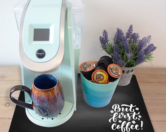 May include: A black chalkboard style mat with the text "But first, coffee!" in white cursive lettering. A teal blue mug with a brown and black design is on the mat next to a light blue coffee maker. A teal blue cup filled with coffee pods is on the mat.