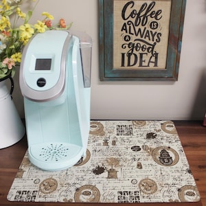Puede incluir: Una cafetera azul claro con una pantalla digital se encuentra sobre una alfombra de cocina beige y marrón con temática de café. La alfombra tiene un diseño vintage con granos de café, tazas de café y texto. Un letrero enmarcado sobre la cafetera dice "El café siempre es una buena idea."