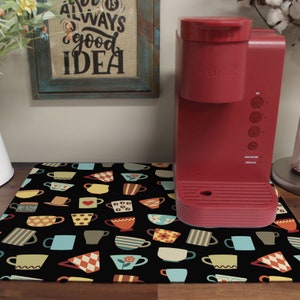 May include: A black and white kitchen mat with a repeating pattern of colorful teacups and saucers. A red single-serve coffee maker is sitting on the mat.