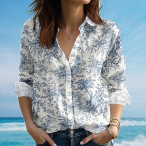 May include: White button-down shirt with a blue toile pattern featuring pastoral scenes. The long-sleeved shirt has a V-neck and is worn with blue jeans. The cuffs are rolled up, and the shirt is unbuttoned at the top.