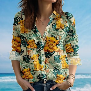 Puede incluir: Camisa con botones color crema con estampado tropical de cachorros de león amarillos y hojas verdes. La camisa de manga larga tiene cuello en V y se usa con jeans azules.