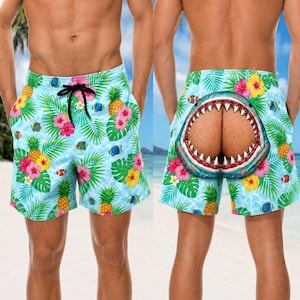 May include: Light blue swim shorts featuring a tropical pattern of yellow pineapples, pink and yellow hibiscus flowers, and small orange and blue fish. The front has a black drawstring, and the back displays a large graphic of a shark mouth.