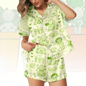 Peut inclure: Ensemble de pyjama vert clair avec un motif répété de personnages de dessins animés, de cœurs et d'étoiles. Le haut à manches courtes et le short ont une bordure blanche et le mot "SMISKI" imprimé. Le tissu a un aspect satiné.