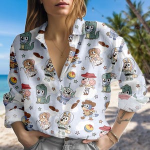 May include: Light blue button-down shirt featuring a repeating pattern of cartoon dogs in various costumes and poses, with stars and other small graphics. The shirt has long sleeves and a V-neck collar.