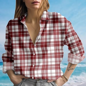 Puede incluir: Una camisa a cuadros rojos y blancos con mangas largas y cuello. La camisa está parcialmente desabrochada. El fondo muestra una escena de playa con agua y cielo azules.