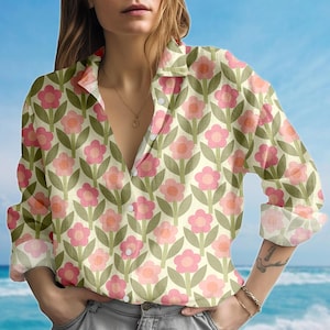 May include: A button-down shirt with a floral pattern. The shirt features a repeating design of pink flowers with orange centers and green leaves on a cream background. The long-sleeved shirt has a collared neckline.