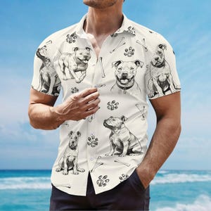 May include: Short-sleeved button-down shirt with a cream background. The shirt features a repeating pattern of black and white illustrations of dogs, paw prints, and bones. The shirt is being worn by a person on a beach.