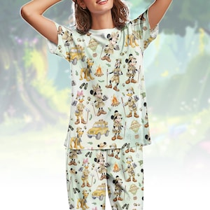 Op de afbeelding: Lichtgroene pyjamaset met een herhalend patroon van stripfiguren in safari-outfits, waaronder Mickey Mouse, Minnie Mouse, Donald Duck en Pluto. Het ontwerp bevat safarivoertuigen, kampvuren en pootafdrukken. De set bevat een top met korte mouwen en een bijpassende broek.