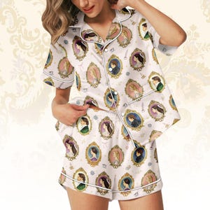 May include: White pyjama set with a repeating pattern of gold and pink framed portraits. The short-sleeved top and shorts are trimmed with white piping. The portraits depict various figures in oval frames, creating a vintage aesthetic.