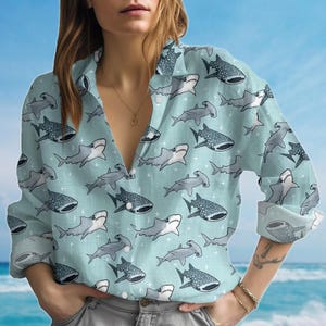 May include: Pale blue button-down shirt with a repeating pattern of grey and white sharks. The shirt has long sleeves and a classic collar. The background is a light blue with a subtle crosshatch pattern.