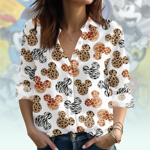 Puede incluir: Camisa blanca con botones con un patrón repetido de siluetas de Mickey Mouse con diseños de estampado de cebra, jirafa y leopardo. La camisa de manga larga tiene cuello en V y se usa con jeans azules.