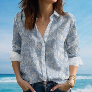 May include: A light blue button-down shirt with a seashell pattern. The long-sleeved shirt features a white seashell design and white cuffs. The shirt is unbuttoned at the top and is paired with blue jeans.