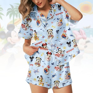 May include: Light blue pajama set featuring a nautical-themed print with cartoon characters. The short-sleeved top and shorts have white trim and a button-down front. The print includes Mickey Mouse, Minnie Mouse, Donald Duck, and other characters in sailor outfits.