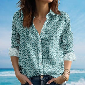 Pode incluir: Uma camisa abotoada com um padrão em forma de leque em verde-azulado e branco. A camisa de manga comprida tem punhos enrolados e decote em V. A camisa é combinada com jeans azuis. O fundo é um céu azul.