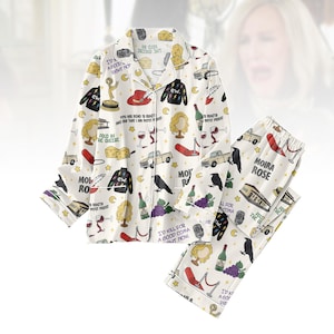 May include: White pajama set with a repeating pattern of cartoon illustrations. The design includes images of wine bottles, crows, and the text "Moira Rose". The set features a button-down shirt and matching pants.