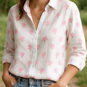 Pode incluir: Camisa branca com estampa floral rosa, combinada com jeans azuis. A camisa de manga comprida tem botões. O padrão floral repete-se na camisa. Um colar de ouro é visível.