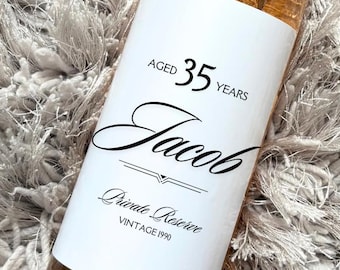 May include: A bottle of rosé wine with a white label featuring the name "Jacob" and the text "Aged 35 Years, Private Reserve, Vintage 1990." The bottle has a silver cap and is set against a fluffy, light gray background.