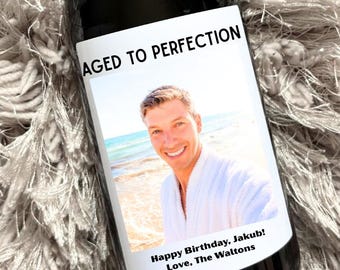 May include: A dark wine bottle with a gold-colored top and a white label. The label reads "AGED TO PERFECTION" above a photo of a man. Below the photo, it says "Happy Birthday, Jakub! Love, The Waltons."