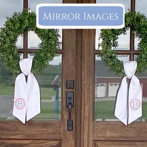 Pu&ograve; includere: Due ghirlande verdi adornano una porta di legno, ciascuna con una fascia bianca. Le fasce sono legate con un fiocco e presentano una lettera "B" monogrammata rosa all'interno di un disegno circolare. Il testo "MIRROR IMAGES" &egrave; visualizzato in un rettangolo blu sopra la porta.