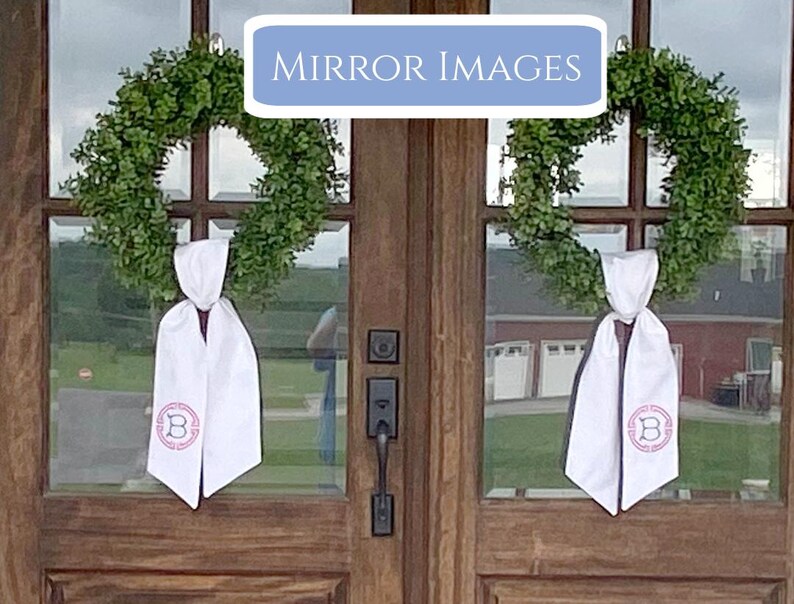 Pu&ograve; includere: Due ghirlande verdi adornano una porta di legno, ciascuna con una fascia bianca. Le fasce sono legate con un fiocco e presentano una lettera "B" monogrammata rosa all'interno di un disegno circolare. Il testo "MIRROR IMAGES" &egrave; visualizzato in un rettangolo blu sopra la porta.