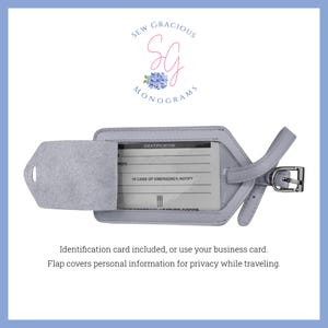 May include: A light grey leather luggage tag with a flap that covers a clear plastic window. The window has a form for identification and emergency contact information. The tag has a strap and a metal buckle.